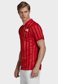 Camisa atlética roja con un patrón de rayas verticales, mangas cortas y un cuello con solapa. Presenta el logo de Adidas en el pecho izquierdo.