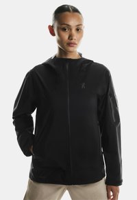 Czarna wodoodporna kurtka z kapturem, zapinana na suwak z przodu oraz z kieszeniami po bokach. Posiada logo na torsie i regulowany dół dla lepszego dopasowania.