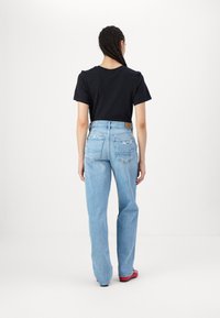 Melna kokvilnas T-krekla un gaiši zilas augsta vidukļa džinsas ar aizmugurējām kabatām un nolietotiem izsistiem elementiem, kopā ar sarkaniem apaviem, skatīts no aizmugures.