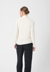 Langärmliger, cremefarbener Rollkragenpullover aus weichem Stoff, mit taillierter Form und glatter Textur. Kombiniert mit schwarzen, maßgeschneiderten Hosen.