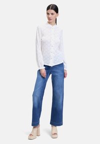 Blusa bianca con bottoni e un motivo in pizzo testurizzato, abbinata a jeans blu a gamba larga e sandali con piattaforma beige.