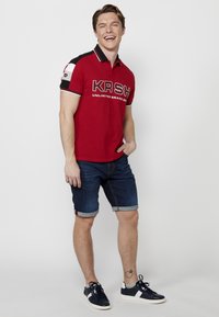 Piros póló fekete kiegészítőkkel és fehér pipingszalaggal, rajta a "KRSH" logóval. Hozzá illő, feltekert farmernadrág. A cipő sötétkék, fehér részletekkel.