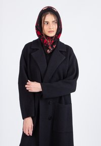 Vrouw in een zwart oversized jasje en een zwarte kapscarf met een rood en roze bloemenpatroon, kijkend naar de zijkant tegen een witte achtergrond.