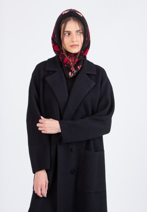 Donna che indossa un cappotto oversize nero e una sciarpa con cappuccio nera decorata con un motivo floreale rosso e rosa, mentre guarda di lato su uno sfondo bianco.