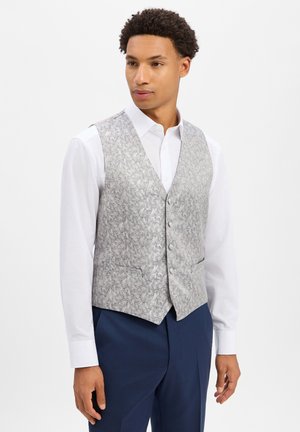 Mann, der eine silberne gemusterte Weste über einem weißen Hemd und marineblaue Hose trägt, steht vor einem einfarbigen hellen Hintergrund.