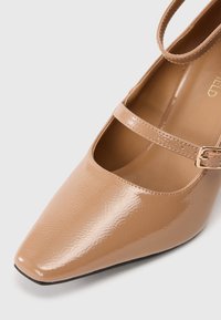 Nude lakovaná Mary Jane bota s špičatou špičkou, disponující jedním páskem a detaily s přezkou. Hladký povrch s mírným leskem.