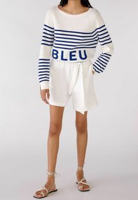 Pull en maille blanche avec des rayures bleues et le texte "BLEU", associé à un short blanc noué à la taille. Le mannequin porte des sandales à talons beige.