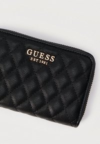 Μαύρο πορτοφόλι με καπιτονέ υφή και ανάγλυφη επιφάνεια, με χρυσό λογότυπο "GUESS EST 1981". Κλείνει με φερμουάρ και έχει αντίθετες ραφές ως διακοσμητικά στοιχεία.