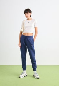 Ung kvinne står på grønn gulv iført hvit crop topp, blå sweatpants, hvite sneakers og holder en kremfarget håndveske på skulderen.