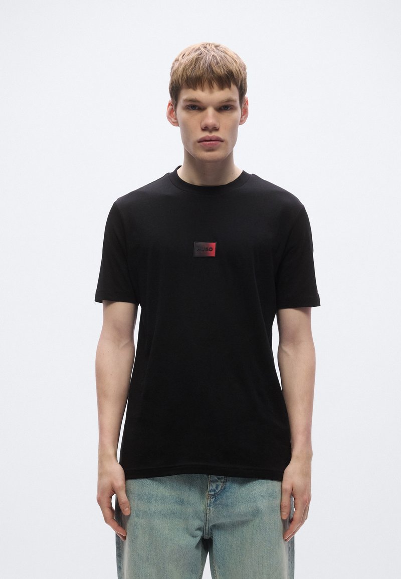 Μαύρο βαμβακερό t-shirt με στρογγυλή λαιμόκοψη, διακοσμημένο με ένα μικρό κόκκινο και μαύρο λογότυπο στο στήθος. Στυλιζαρισμένο με ανοιχτόχρωμο τζιν παντελόνι.