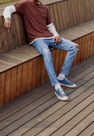 Lässiges Outfit mit einem weinroten T-Shirt über einem weißen Langarmshirt, hellblauen Zerrissenen Jeans und mehrfarbigen gemusterten Sneakers.