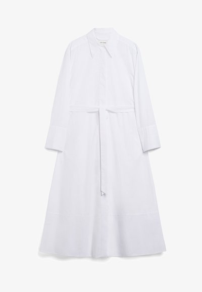 Lungo vestito camicia bianco realizzato in cotone. Presenta un colletto a punta, maniche lunghe, una vita con fiocco frontale e un sottile dettaglio all'orlo.
