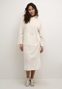 Cremefarbenes Hoodie-Kleid mit einer Kängurutasche, Kapuze mit Kordelzug, langen Ärmeln und einer Länge bis zur Mitte der Wade. Kombiniert mit grauen und beigen Sneakers.