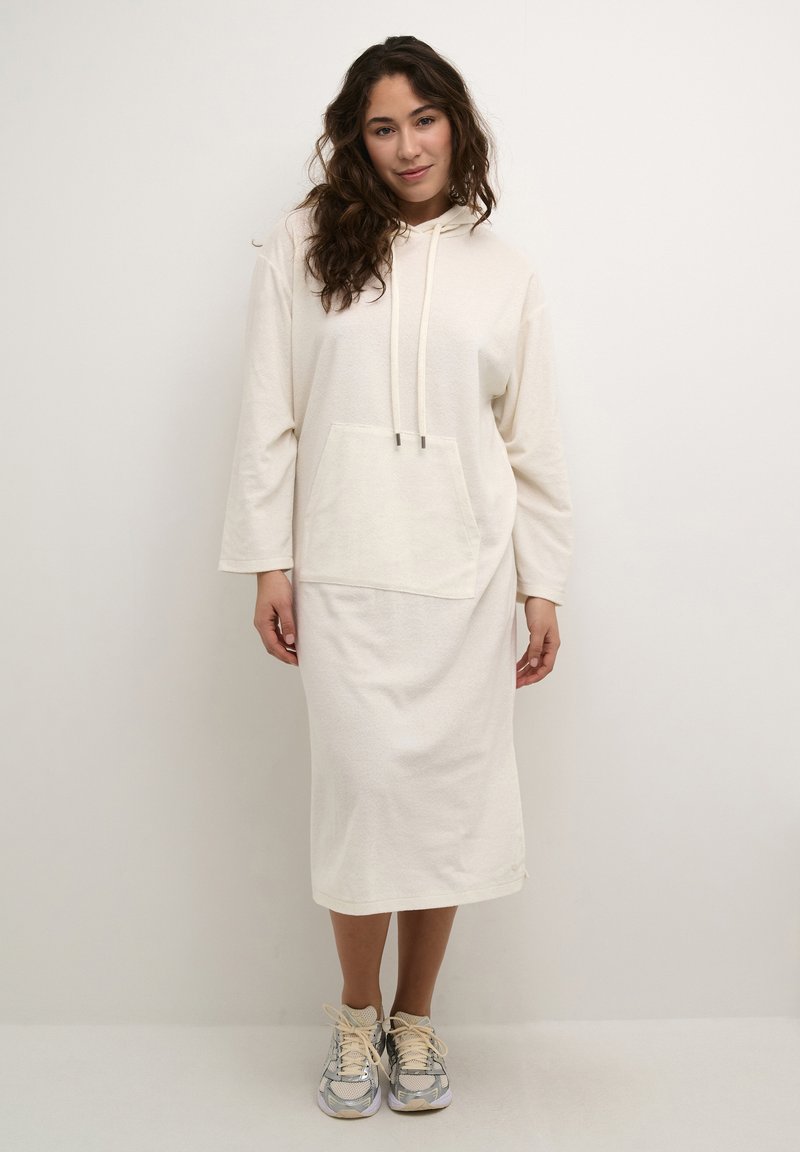Cremefarbenes Hoodie-Kleid mit einer Kängurutasche, Kapuze mit Kordelzug, langen Ärmeln und einer Länge bis zur Mitte der Wade. Kombiniert mit grauen und beigen Sneakers.