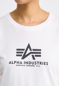 T-shirt blanc à manches courtes arborant un logo graphique noir avec des ailes stylisées et le texte "ALPHA INDUSTRIES". Matière en coton douce et légère.