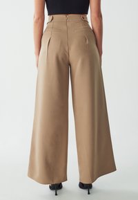 High-waisted, wide-leg trousers in beige fabric. Features include back pockets, tailored pleats, and a smooth, textured finish.