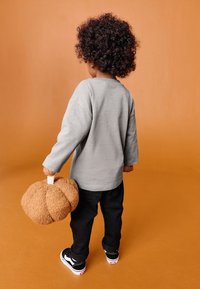 Szara koszula z długim rękawem, czarne spodnie i czarne trampki. Trzyma teksturowaną brązową torebkę w kształcie dyni z białym uchwytem na pomarańczowym tle.