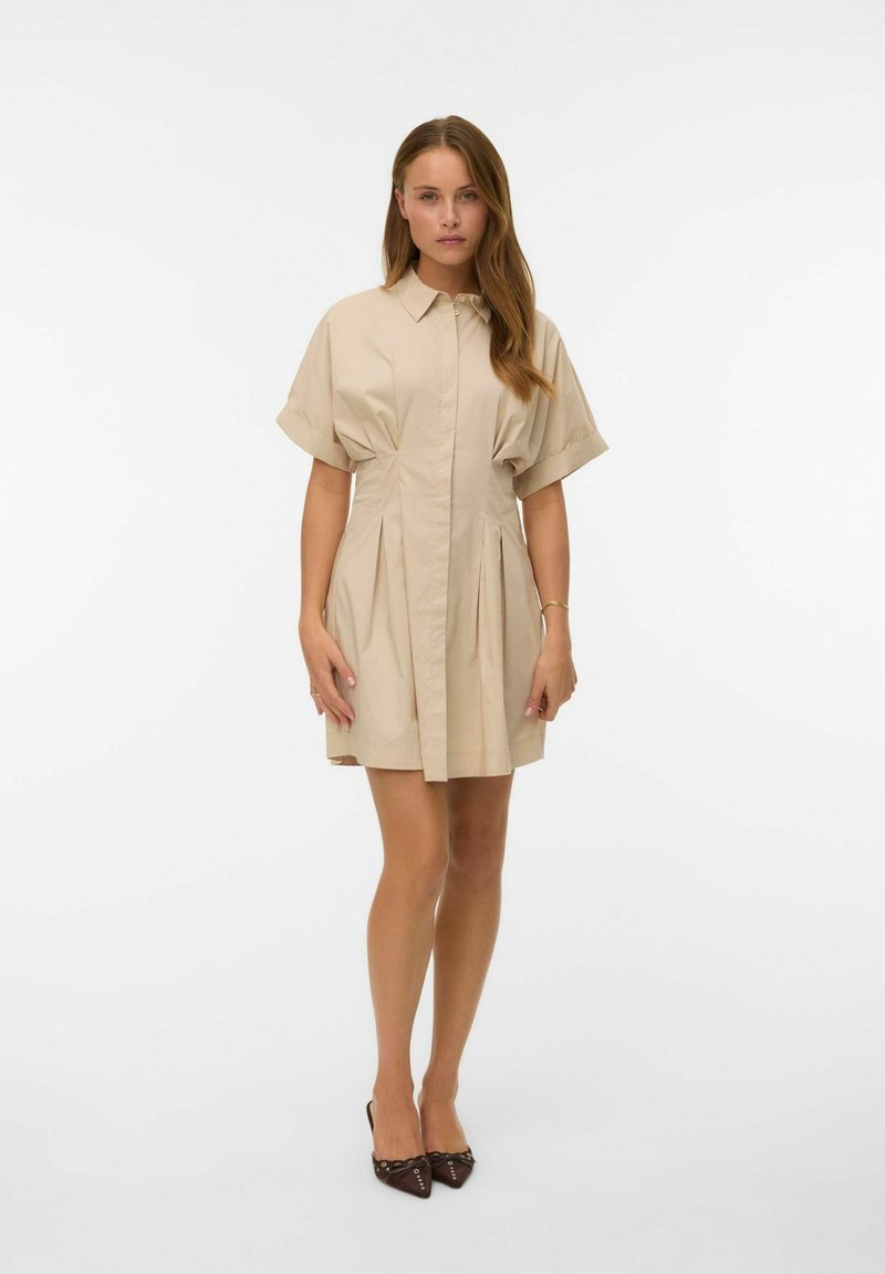 Femme portant une robe beige à manches courtes, col avec boutons et plis, debout devant un fond blanc uni, chaussures noires pointues.