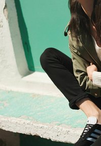 Czarne trampki z niebieskimi i białymi paskami, czarne wąskie dżinsy oraz oliwkowa kurtka na białym swetrze, siedzący na teksturowanej turkusowej powierzchni.