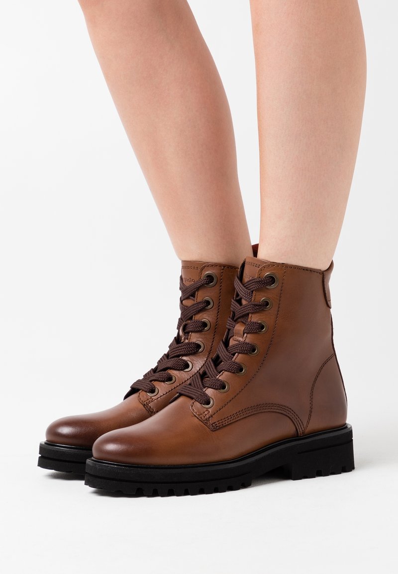 Bottes courtes en cuir marron avec des lacets marron foncé, tige texturée, bout rond et semelle en caoutchouc noire robuste, conçues pour la durabilité et le soutien.