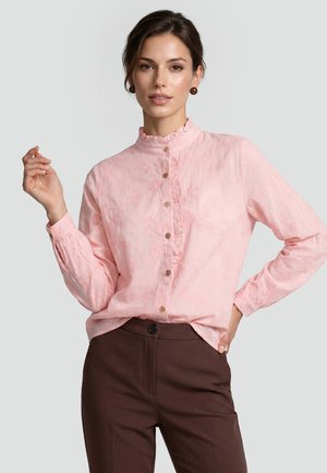 Frau trägt eine hellrosa Bluse mit Knöpfen und Blumenstickerei und dunkelbraune, hoch taillierte Hose, posiert mit einer Hand in der Taille.