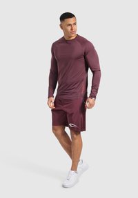 Maglietta atletica a maniche lunghe bordeaux con maniche raglan, abbinata a pantaloni corti coordinati con un logo laterale, completata da sneakers bianche.