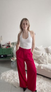 Νεαρή γυναίκα στέκεται με τα χέρια στις τσέπες, φοράει λευκό τοπ με λουρί στο λαιμό και ψηλόμεσο, φαρδύ, φωτεινό ροζ παντελόνι σε ένα ζεστό καθιστικό.