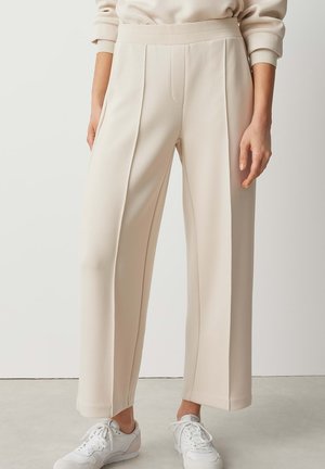 Pantalon ample couleur crème avec plis sur le devant, porté avec des baskets blanches et un sweat-shirt beige clair, montré de la taille aux pieds.