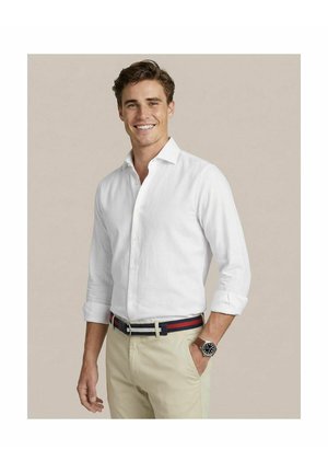 Jonge man die lacht, een wit overhemd met knopen draagt, beige broek, een gestreepte riem en een horloge, met één hand in de zak.
