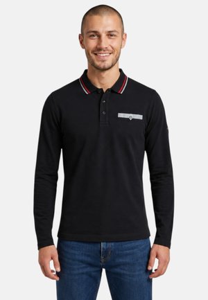 Jeune homme souriant portant un polo noir à manches longues avec des rayures rouges et blanches au col et un jean bleu sur fond blanc.