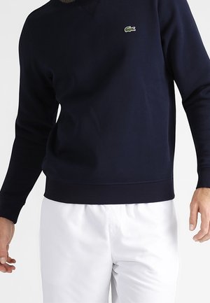 Homme portant une chemise bleu marine à manches longues avec un petit logo crocodile vert et un pantalon blanc sur un fond uni.