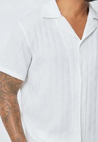 Chemise blanche à manches courtes avec un motif texturé, dotée d'un col en V et de boutons, fabriquée en tissu léger.
