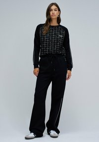 Femme debout portant un sweat à manches longues à carreaux noirs, un pantalon noir ample avec des rayures sur les côtés, et des baskets blanches sur fond gris.