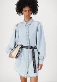 Gaiši zils denim kleitas krekls ar pogu priekšpusi, pūkainām piedurknēm un melnu logo jostu. Tur rokā mazu sudraba rokassomu.
