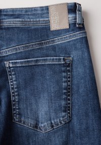 Vaqueros de mezclilla azul oscura con un bolsillo trasero cosido, tejido texturizado y una etiqueta de cuero gris claro en la cintura con texto en relieve.