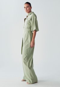 Moteris stovi šalies žaliai spalvotame kombinezone su pūstomis rankovėmis, plačiomis kojomis ir juosmens raiščiu, žiūrėdama į kamerą prieš paprastą foną.