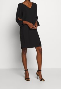 Czarna sukienka z dekoltem w serek i długimi, otwartymi rękawami oraz bocznymi rozcięciami. Gładki materiał o luźnym kroju. Połączona z czarnymi sandałami na wysokim obcasie z paskami.
