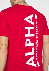 Crvena pamučna majica s velikim bijelim printom "ALPHA INDUSTRIES" okomito uz lijevu stranu. Kratki rukavi, dizajn s okruglim izrezom.
