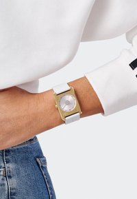 Montre rectangulaire dorée avec cadran argenté et bracelet en silicone blanc, arborant un design minimaliste et de petits repères horaires en argent.