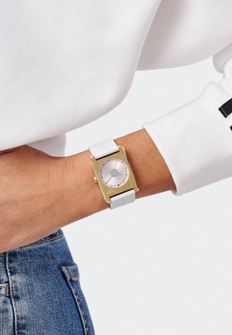 Gold rectangular watch with silver dial and white silicone strap, featuring a minimalist design and small silver hour markers.