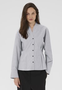 Kvinne med langt brunt hår iført en lysegrå knappet bluse og sorte bukser foran en ensfarvet hvid baggrund.