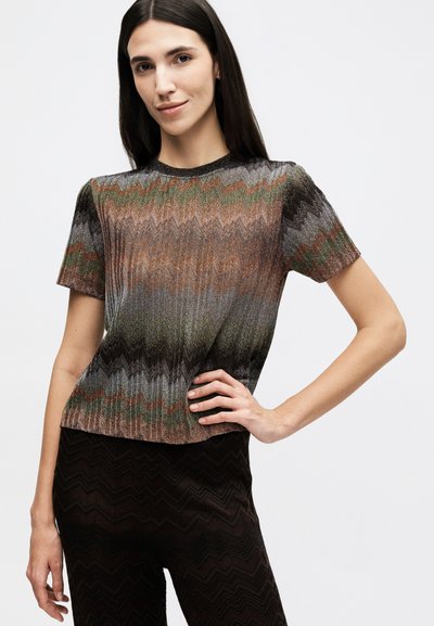 Žena s dlouhými tmavými vlasy, oblečená v krátkém rukávu s vícero barevným vzorem cik-cak na topu a černé kalhoty se vzorem cik-cak, pózující s rukou na bocích.