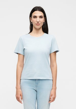 Femme aux cheveux raides et foncés, portant un t-shirt bleu clair à manches courtes et un jean bleu clair, debout devant un fond blanc uni.