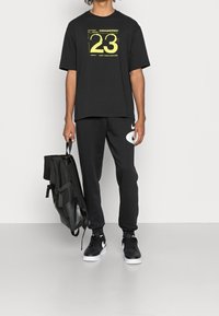 Černé tričko s krátkým rukávem a žlutou grafikou „23“, spárované s černými tepláky s bílým logem Nike. Drží černý batoh.
