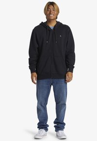 Sort zip-hoodie lavet af fleece, med en hætte med snoretræk, forlommer og ribbede manchetter, parret med blå denimjeans og sneakers.