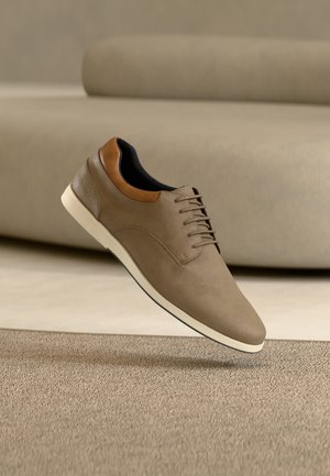 Chaussure décontractée marron à lacets avec semelle blanche et col beige flottant au-dessus d'un tapis beige devant un canapé assorti.