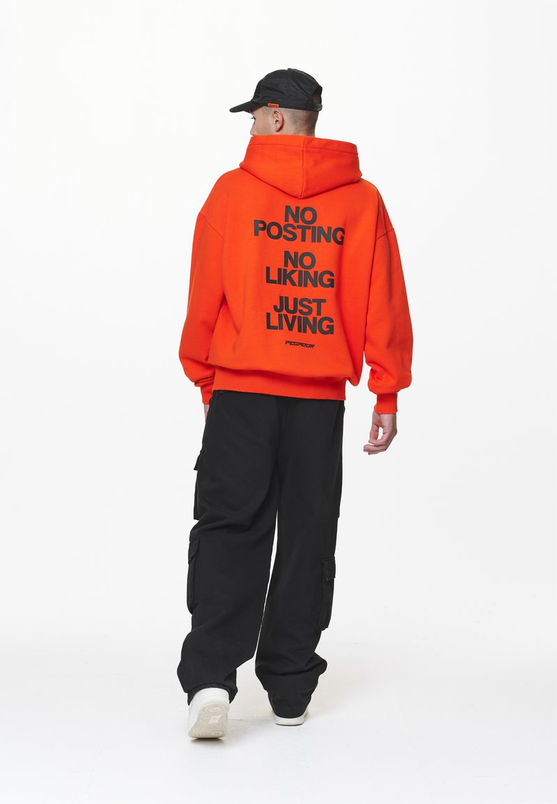 Hanorac portocaliu cu text negru: "FĂRĂ POSTĂRI, FĂRĂ APRECIERI, DOAR TRĂIRE." Asortat cu pantaloni cargo negri, largi, și adidași albi. Şapcă neagră purtată pe dos.