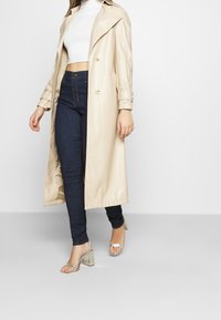 Trench-coat beige texturé avec un design double boutonnage et de grands revers. Porté par-dessus un jean foncé et des sandales à talons blocs transparentes.