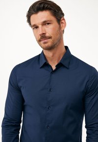Camicia blu navy con bottoni, colletto, maniche lunghe e una texture liscia. Presenta bottoni piccoli e un design aderente.