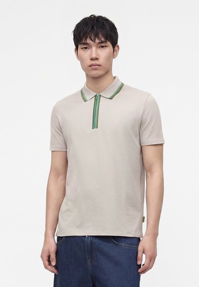 Jeune homme portant un polo beige à manches courtes avec un col rayé vert et un jean bleu foncé, debout sur un fond blanc.